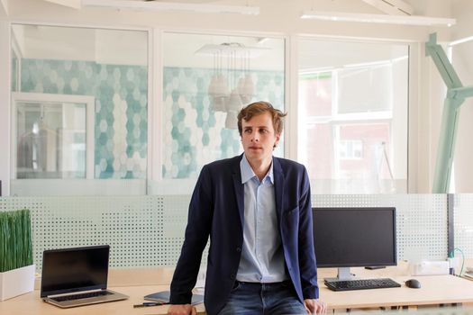 Relaxed modern businessman in suit standing inside of contemporary glass office sitting on table with computers