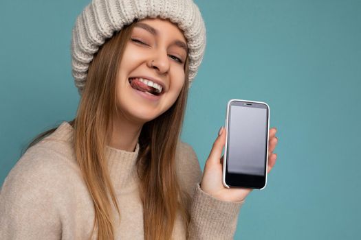 Closeup portrait of beautiful sexy positive good looking young woman poising isolated over blue background with empty space holding in hand and showing mobile phone with empty screen for mockup looking at camera and showing tongue.