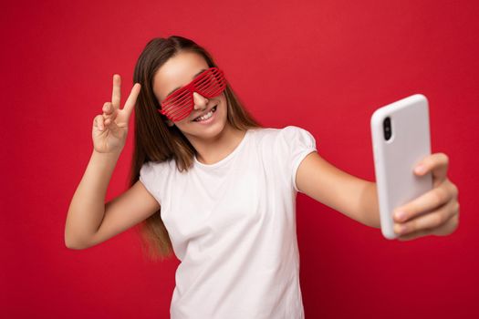 Charming positive young girl wearing white t-shirt and red glasses poising isolated on red background with empty space holding in hand and using mobile phone ctaking selfie looking at gadjet camera and showing peace gesture