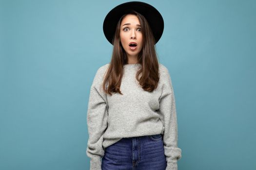 Young beautiful brunet woman. Trendy lady in casual grey sweater and stylish black hat. Positive female shows facial sincere emotions. Funny amazed surprised model isolated on blue background with free space.