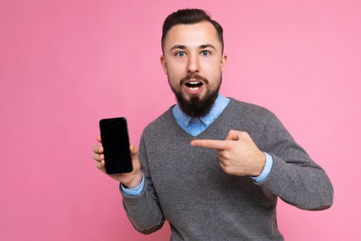 Photo of handsome smiling adult male person good looking wearing casual outfit standing isolated on background with copy space holding smartphone showing phone in hand with empty screen display for mockup pointing at gadjet looking at camera.