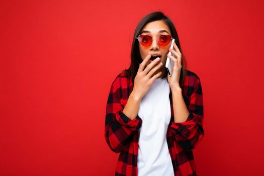 Beautiful shocked amazed young brunette woman wearing stylish red shirt white t-shirt and red sunglasses isolated over red background talking on mobile phone looking at camera.