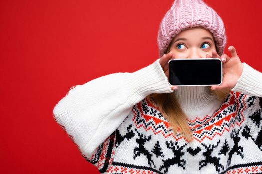 Photo of attractive shocked young blonde woman wearing warm knitted hat and winter warm sweater standing isolated over red background showing smartphone with empty screen for cutout looking to the side.