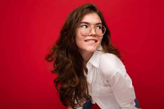 Photo shot of beautiful positive young brunette woman wearing casual clothes and stylish optical glasses isolated over colorful background wall looking to the side.