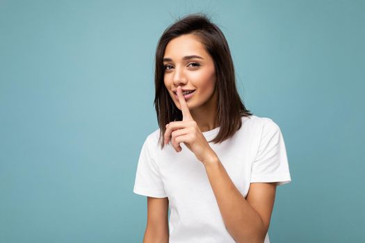 Portrait of young attractive brunette woman with sincere emotions wearing casual white t-shirt for mockup isolated on blue background with copy space and showing shhh gesture. Be quiet. Don't speak.