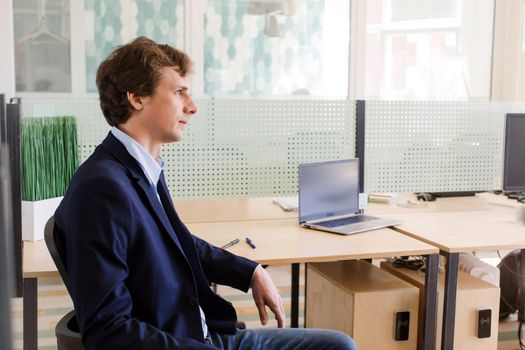 Side view of serious young employee sitting in office near table with laptop