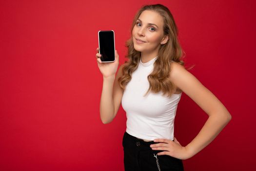 beautiful young blonde woman wearing white t-shirt isolated on red background with copy space holding smartphone showing phone in hand with empty screen for mockup looking at camera.