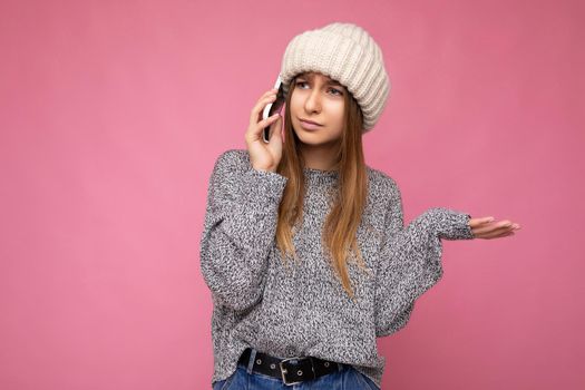 Attractive upset young blonde woman wearing casual grey sweater and beige hat isolated over pink background holding in hand and talking on mobile phone looking to the side and having questions.