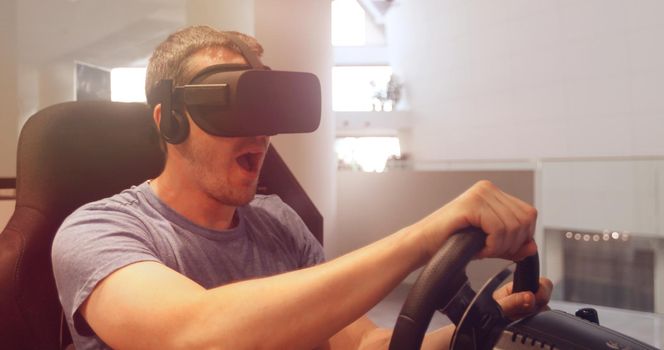 Computer simulation. Cheerful young gamer in virtual reality goggles is expressing excitement while enjoying car racing video game with steering wheel