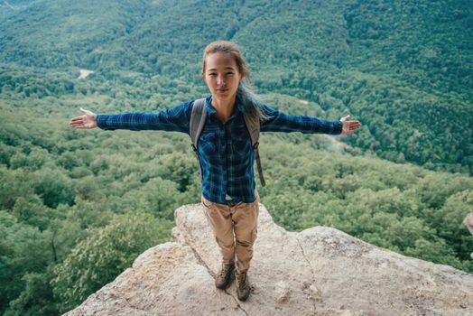 Happy hiker young woman with backpack standing with raised arms in summer mountains, looking at camera