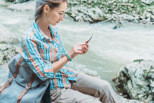 Traveler young woman with backpack sitting on the river bank and using smartphone outdoor