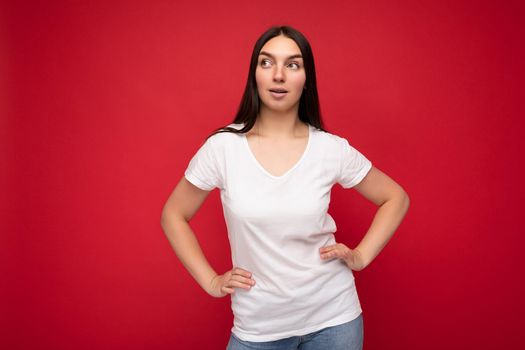Portrait of positive thoughtful young attractive brunette woman with sincere emotions wearing casual white t-shirt for mockup isolated over red background with copy space.