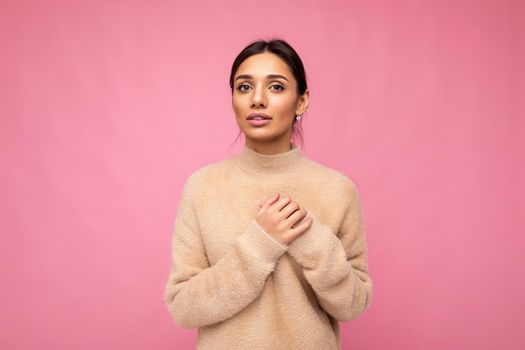 Portrait of young positive beautiful brunette woman with sincere emotions wearing beige pullover isolated over pink background with copy space and holding hand on chest.