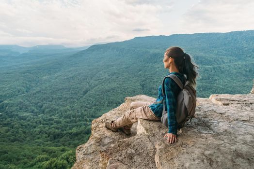 Traveler young woman sitting on edge of cliff and looking on mountains in summer