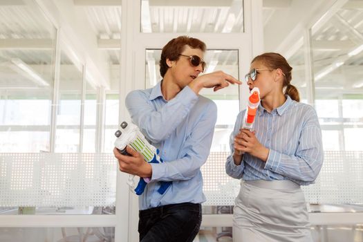 Stylish modern woman and man in sunglasses having fun in office with toy plastic guns