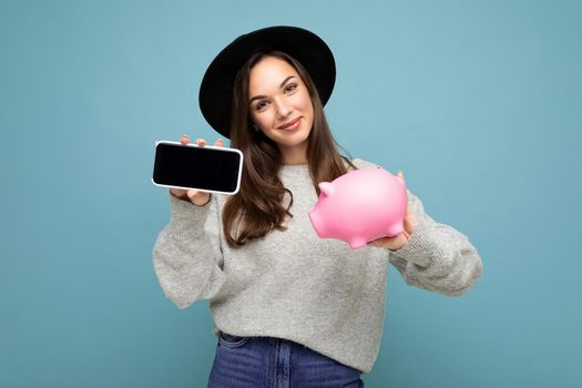 Shot of happy positive smiling satisfied sincere young pretty nice woman with dark hair wearing stylish clothes and black hat isolated on blue wall with copy space and holding pink pig money box and showing telephone with empty screen display.