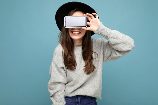 Pretty young smiling woman wearing black hat and grey sweater holding phone looking at camera isolated on background.Mock up, cutout, empty space