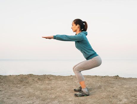 Fitness young woman doing squat exercises on coast outdoor, side view