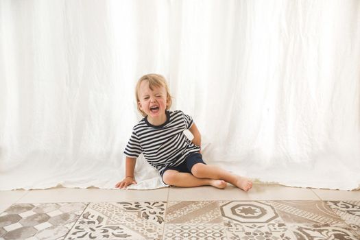 Little adorable boy sitting on floor against white curtain and crying heavily with grimace