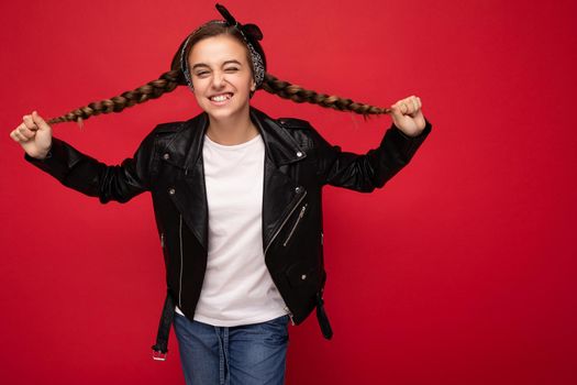 Photo shot of beautiful happy smiling brunette little girl with pigtails wearing trendy black leather jacket and white t-shirt for mockup standing isolated over red background wall looking at camera.