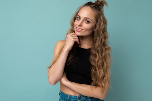 Young beautiful curly blonde woman with sexy expression, cheerful and happy face wearing trendy black top isolated over blue background with copy space.