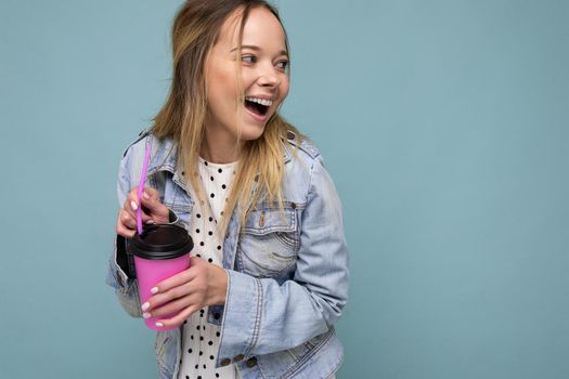 Charming young happy blonde woman wearing stylish clothes isolated over colourful background wall holding paper cup for mockup drinking milkshake looking to the side. copy space