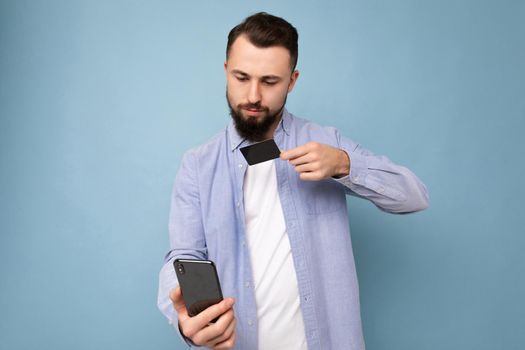 Photo of good looking attractive brunet unshaven young man wearing casual stylish blue shirt and white t-shirt isolated over blue background wall with copy space for text holding black credit card and making selfie on mobile phone.