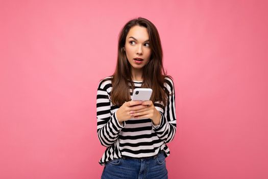 shot of pretty young brunette woman using mobile phone communicating via texting sms wearing sweater isolated on wall background looking to the side.