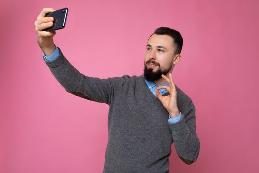 Handsome young man wearing casual stylish clothes standing isolated over background wall holding smartphone taking selfie photo looking at mobile phone screen display.