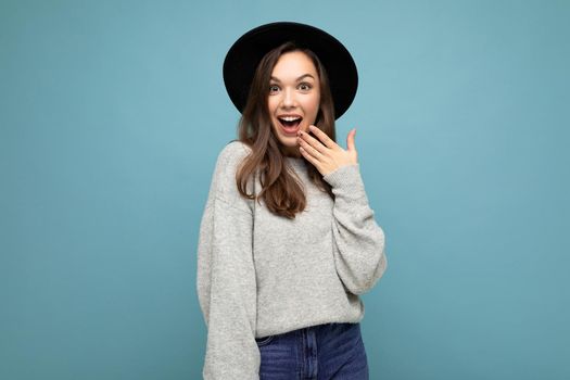 Photo of attractive charming brunette happy amazed shocked young woman wearing grey jersey and black hat isolated on blue background with copy space.
