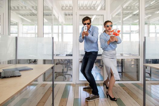 Man and woman in formal outfits standing back to back and aiming toy guns while having fun in office