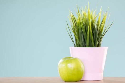 Delicious green apple lying on light blue background near ceramic pot with nice plant