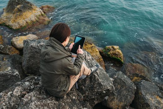 Young woman sitting on stone near the sea and working on digital tablet