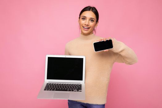 Photo of charming pretty beautiful smiling young woman with dark hair looking at camera holding computer laptop and mobile phone with empty monitor screen with mock up and copy space wearing light colour sweater isolated on pink wall background.