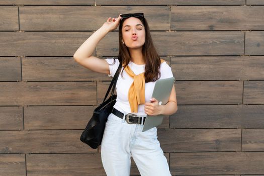 Beautiful charming young brunet woman looking at camera holding computer laptop and sunglasses showing peace gesture with kissing lips and close eyes in white t-shirt and light blue jeans in the street.