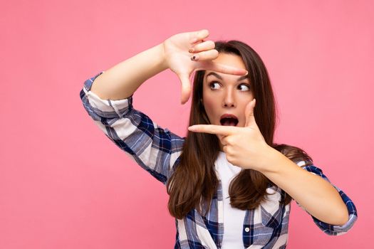 Young positive delightful amazed pretty brunette woman with sincere emotions wearing hipster check shirt making frame with hands, taking picture with imaginary camera and standing isolated on pink background with copy space.