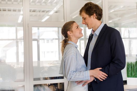 Side view of smiling young male and female colleagues in love hugging and chatting happily during break in modern office building