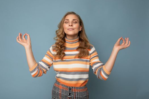 Portrait of young attractive blonde curly woman with sincere emotions wearing trendy striped pullover isolated on blue background with copy space and doing mediation gesture with fingers. Yoga concept.