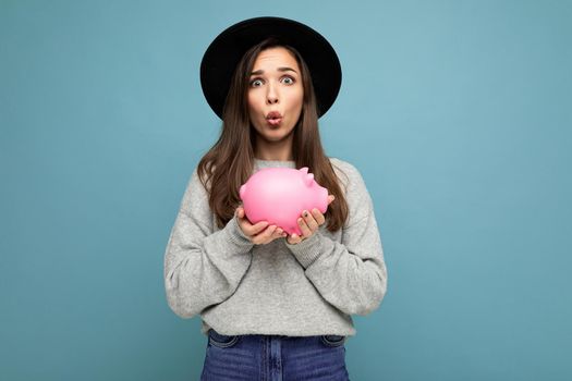 Portrait of shocked amazed funny young pretty charming brunette woman wearing stylish gray sweater and black hat isolated over blue background with empty space and holding pink piggy box. Money box concept.