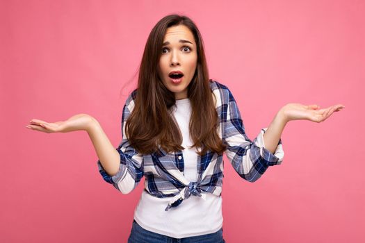 Portrait photo of young surprised astonished beautiful brunette woman with sincere emotions wearing stylish check shirt isolated on pink background with copy space and having doubts.