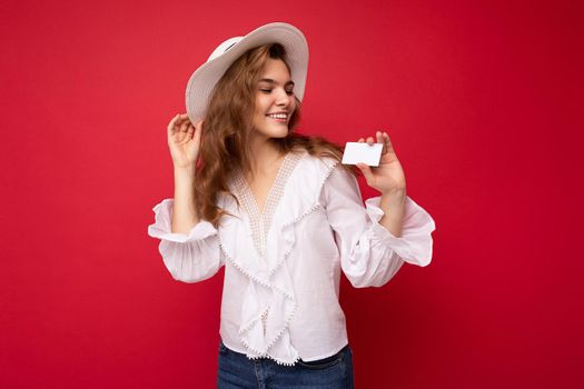 Shot of attractive positive smiling young dark blonde woman wearing white blouse and white hat isolated over red background holding credit card looking at contactless bank card.