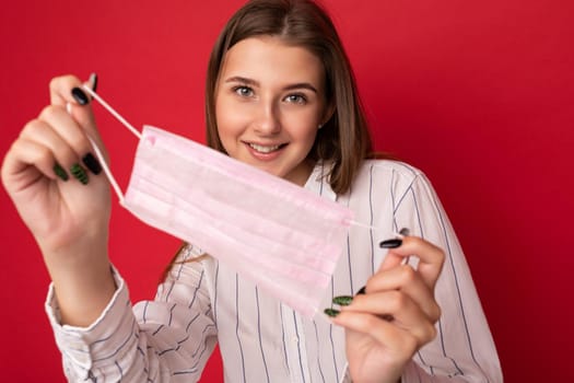 Positive smiling woman holding virus protective mask against coronavirus and wearing white medical coat isolated on red background.