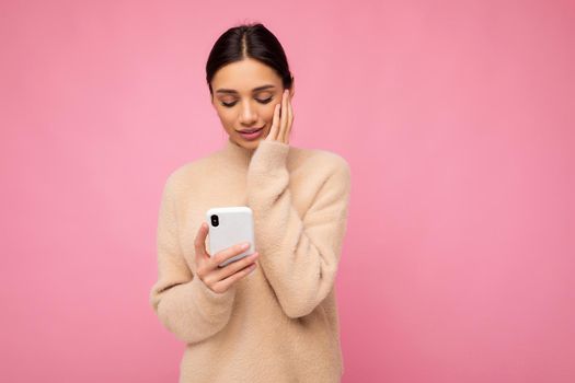 Attractive cute young brunet woman wearing beige warm sweater standing isolated over pink background surfing on the internet via phone looking at mobile screen and being concerned.