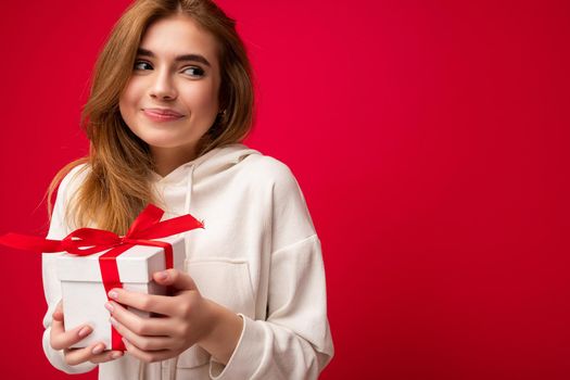 Shot of attractive positive smiling young blonde woman isolated over colourful background wall wearing everyday trendy outfit holding gift box and looking to the side. Empty space
