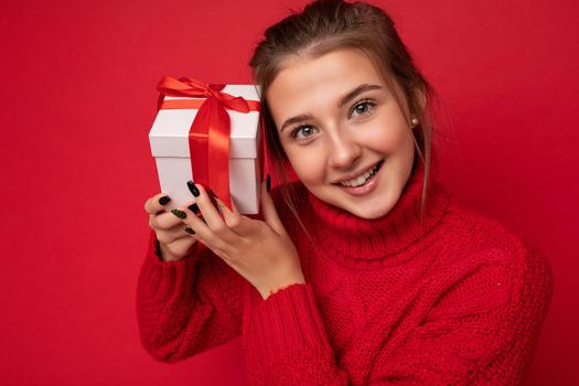 Beautiful happy young brunette woman isolated over colourful background wall wearing stylish casual clothes holding gift box and looking at camera.