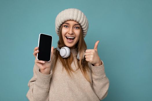 Photo of attractive positive joyful young dark blonde woman wearing beige stylish sweater and knitted beige winter hat isolated on blue background wearing white bluetooth wireless earphones and listening to cool music and showing mobile phone with empty display for mockup looking at camera and showing thumbs up gesture.