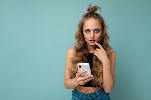Photo of beautiful young blonde woman thoughtful wearing casual stylish clothes holding and using telephone isolated over blue backgrounf looking at camera and thinking.