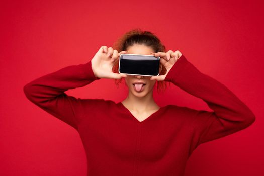 Photo portrait of happy funny young woman wearing dark red sweater isolated over red background holding smartphone and showing mobile phone screen with empty space for mockup having fun.