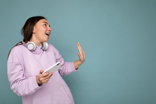 Beautiful happy smiling young brunette woman wearing purple hoodie isolated on blue background holding and using mobile phone surfing on the internet wearing white bluetooth headphones and having fun looking to the side and moving. Copy space