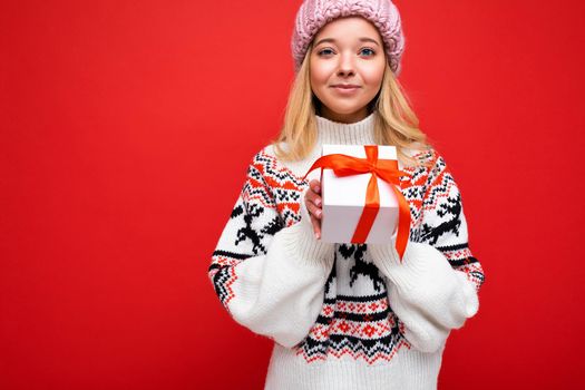 Beautiful happy young blonde woman isolated over colourful background wall wearing stylish casual clothes holding gift box and looking at camera. Copy space, mockup